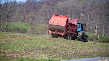 Farm tractor with red manure spreader fertilizes green agricultural field woodland hillside background - stock photo