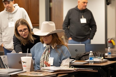 Participant in a white cowboy hat reviews information on a laptop while another attendee points to the screen during a hands-on training session. Other participants work at tables in the background.