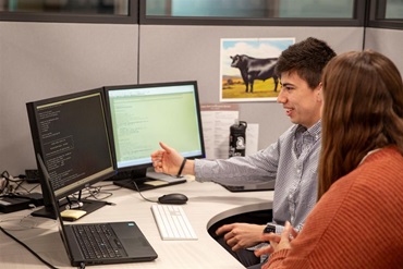 Two individuals discuss genetic evaluation methods at a computer desk in an office.