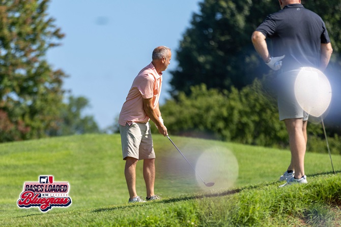 A golfer prepares an approach shot from the edge of the fairway while fellow participants watch during tournament play.