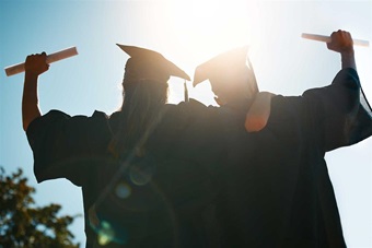 Two graduates raising their hands with their diplomas.