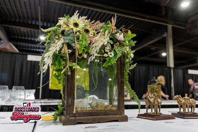 Decorative glass shadow box featuring a cow figurine and floral arrangement displayed at a silent auction.