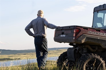 A farmer looking over a large pasture of grass while leaning on a small utility machine.