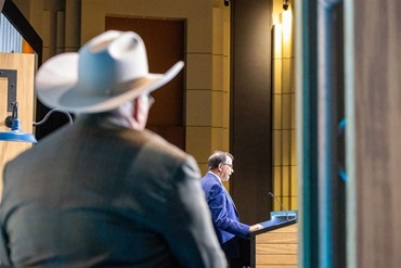 A man in a cowboy hat looks on at a board candidate giving his presentation to the delegate body.