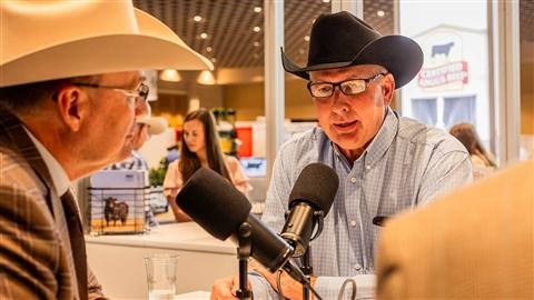 Two men in cowboy hats sit across the table from each other with microphones